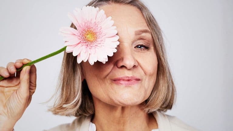 Older woman smiling while holding a pink flower over one eye against a soft neutral background.