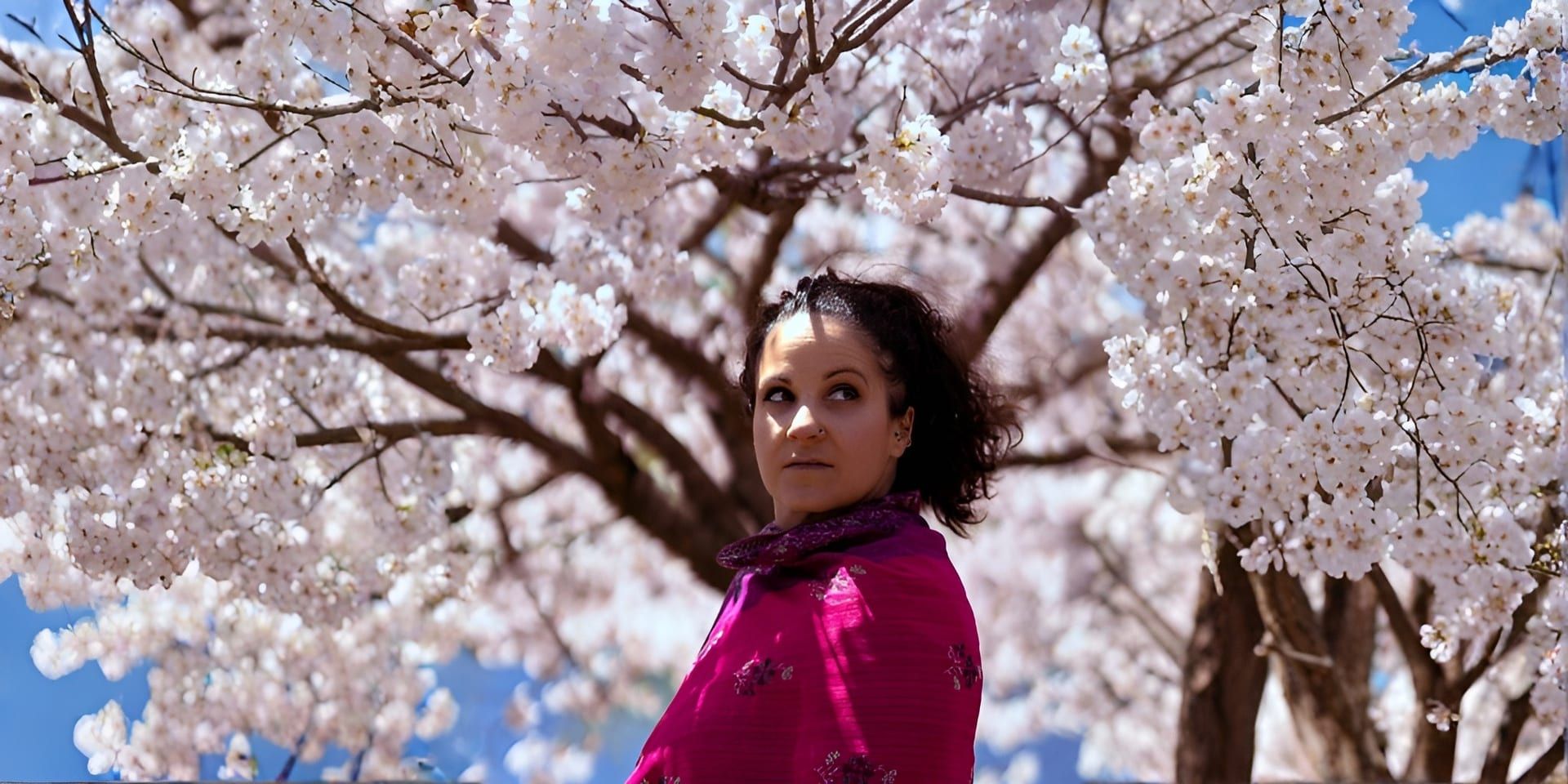 Woman in a vibrant pink shawl stands among blossoming cherry trees, gazing thoughtfully. The blue sky and white blossoms convey a serene, springtime mood.