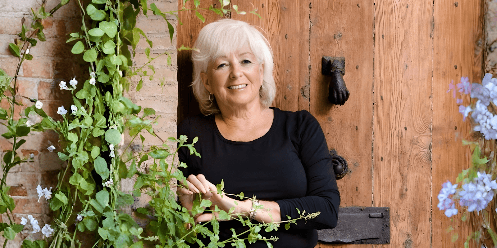 Patricia Sands smiling warmly, surrounded by greenery, leaning against a rustic wooden door.