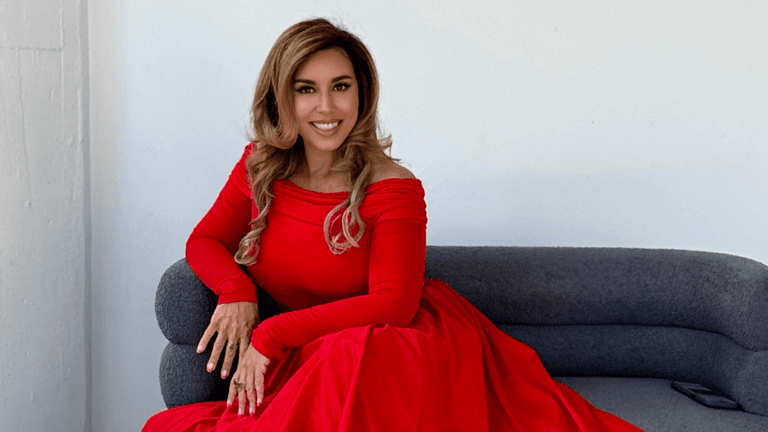 Woman in elegant red off-shoulder gown sitting on gray sofa, smiling against minimalist white wall background.