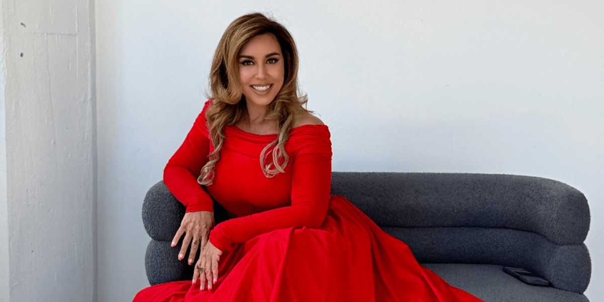 Woman in elegant red off-shoulder gown sitting on gray sofa, smiling against minimalist white wall background.