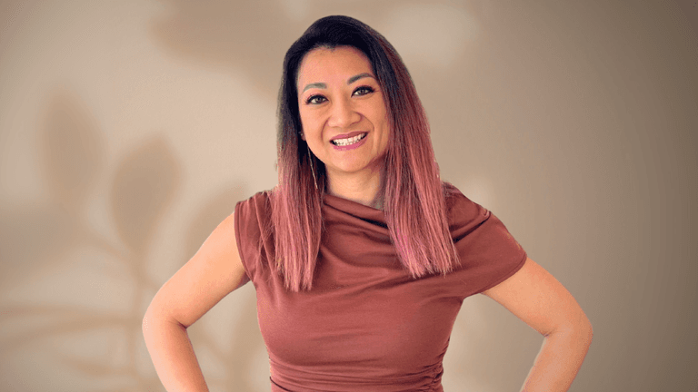"Portrait of a woman with long pink ombre hair, wearing a brown top, smiling confidently with her hands on her hips. The background is a soft, neutral shade with subtle shadows from plants."