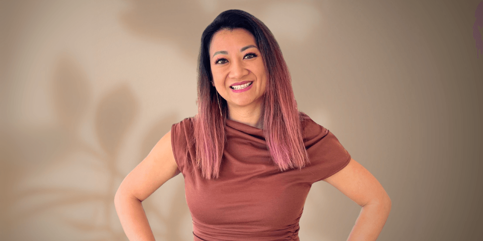 "Portrait of a woman with long pink ombre hair, wearing a brown top, smiling confidently with her hands on her hips. The background is a soft, neutral shade with subtle shadows from plants."