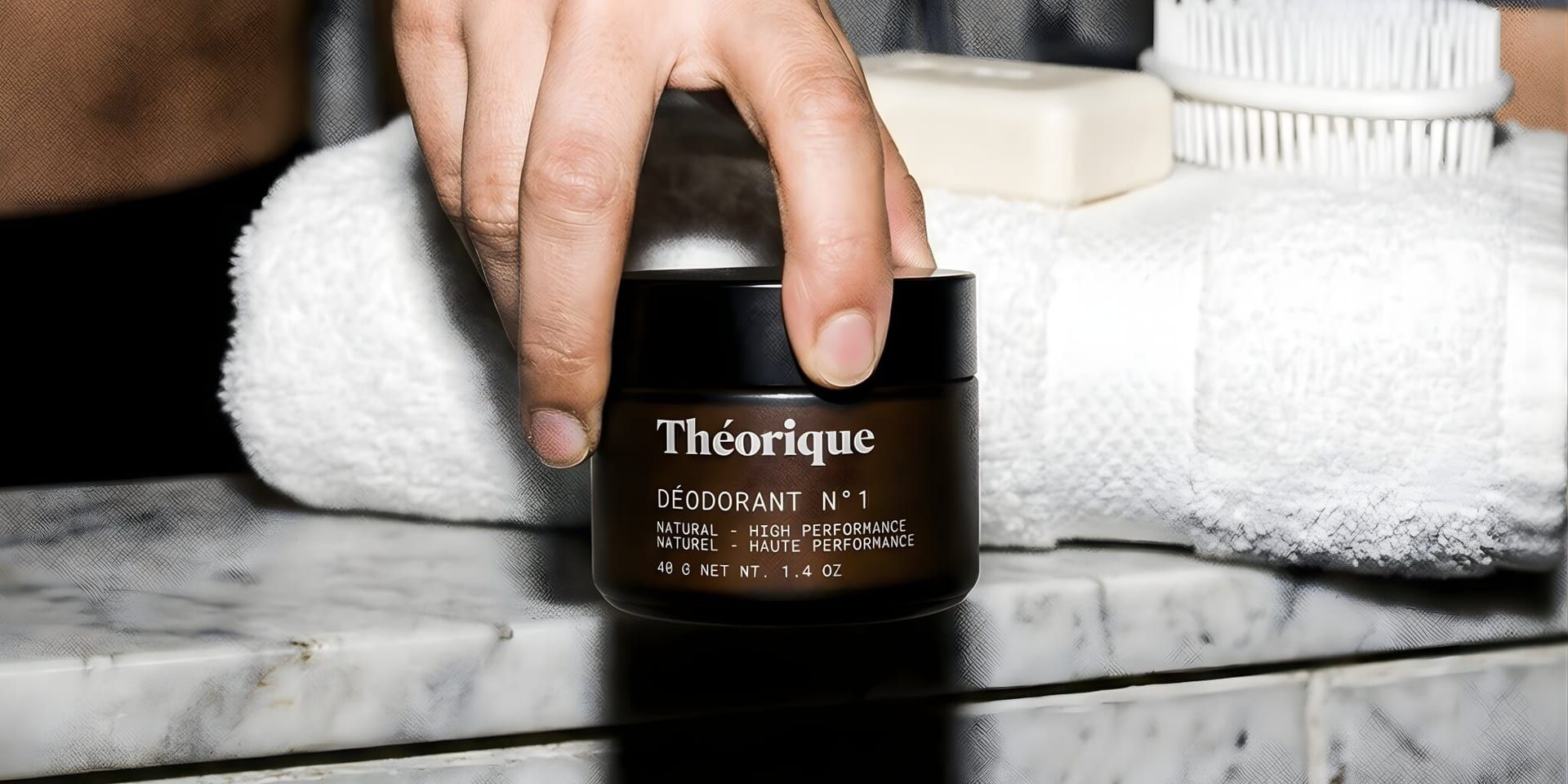 A hand holds Théorique Deodorant N°1 by a rolled white towel, soap, and brush on a marble surface, evoking a clean, minimalist feel.