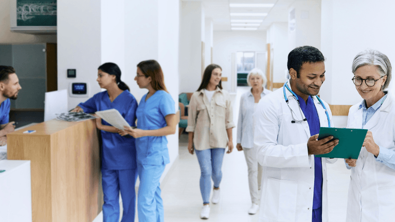 Doctors review a clipboard while nurses and patients stand in a hospital corridor.