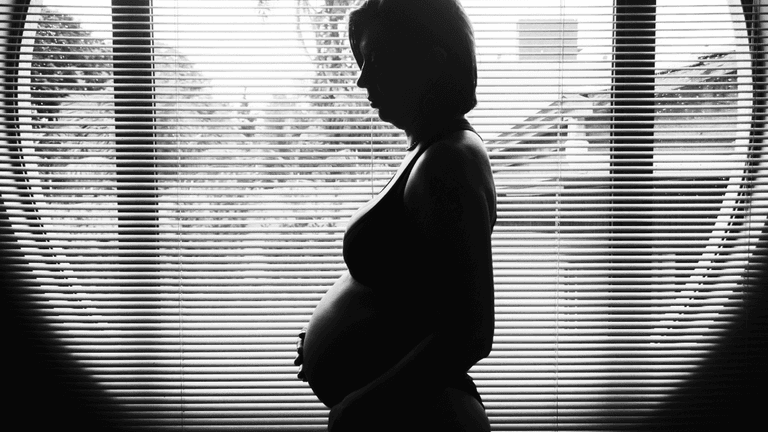 Silhouette of a pregnant woman standing near a window with blinds, creating a peaceful and intimate moment in black and white photography.