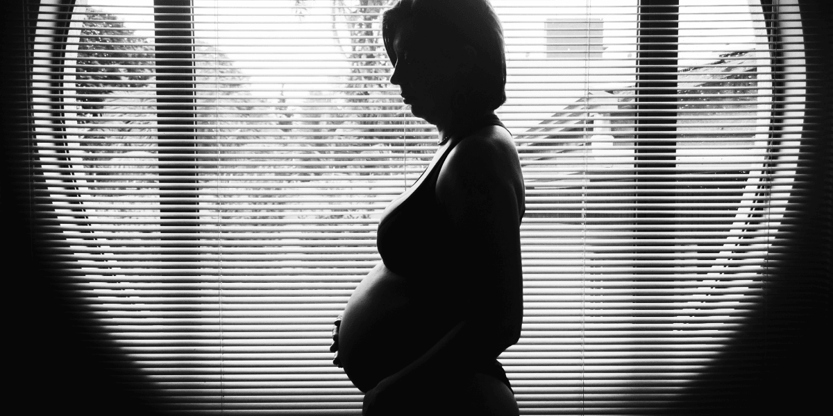 Silhouette of a pregnant woman standing near a window with blinds, creating a peaceful and intimate moment in black and white photography.