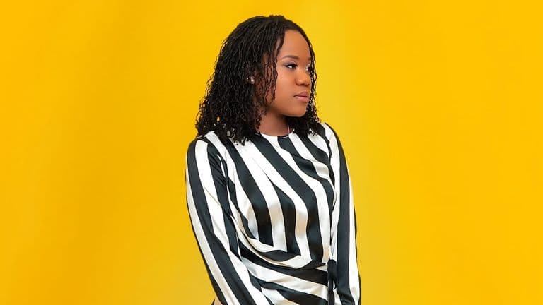 Portrait of a young woman with curly hair wearing a striped black and white top against a vibrant yellow background.
