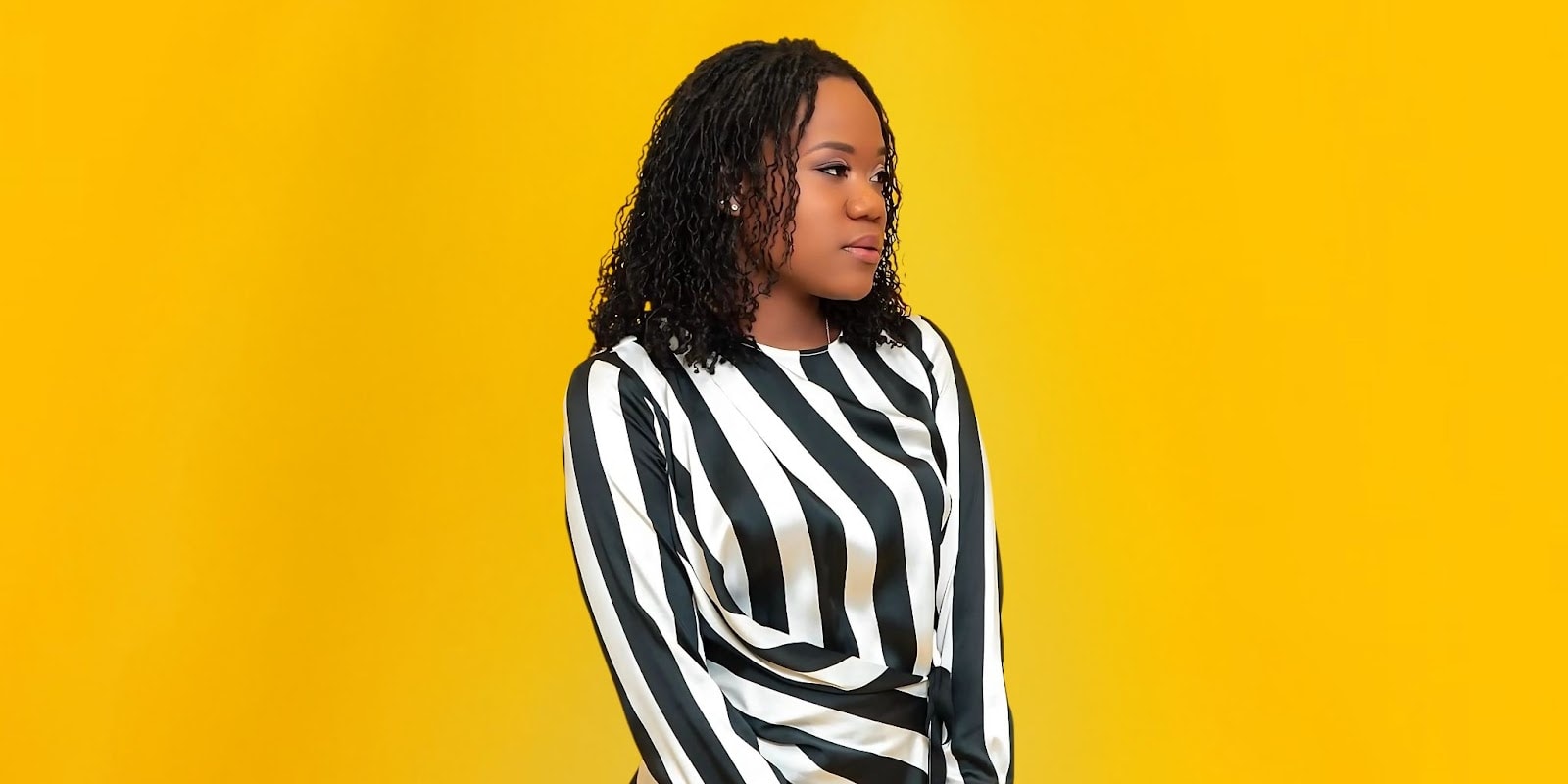 Portrait of a young woman with curly hair wearing a striped black and white top against a vibrant yellow background.