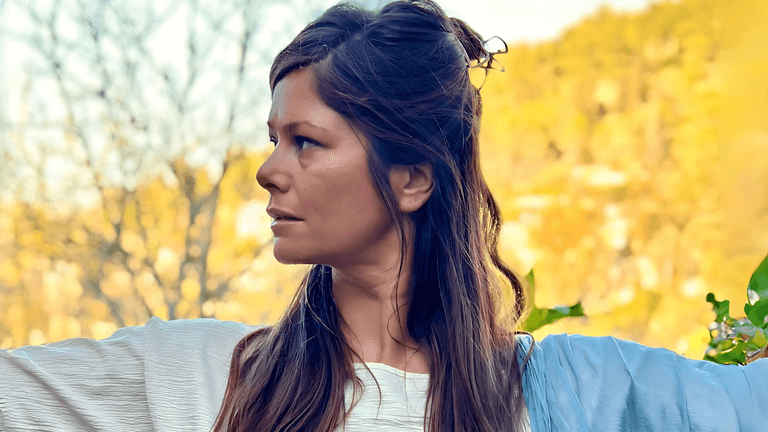A woman stands gracefully outdoors, with a serene and focused expression, embodying the energy of a Tantric Priestess. Her hair is styled in loose waves, and she wears a white robe with a blue sash, symbolizing the harmony of the sacred feminine energy. The warm, natural surroundings of the Galilee mountains reflect the tranquil and transformative environment of Catherine Tiferet Levi's teachings.
