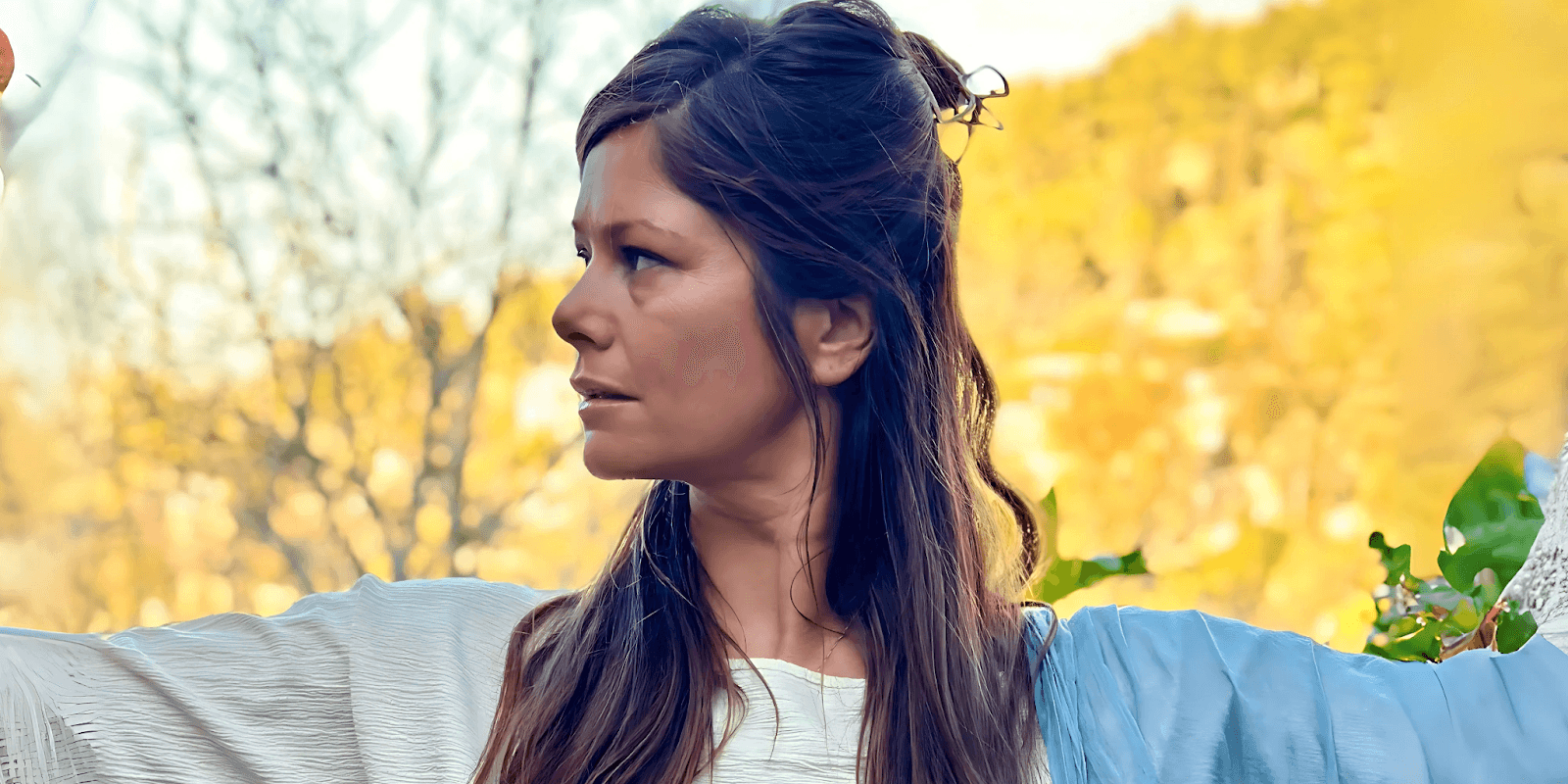 A woman stands gracefully outdoors, with a serene and focused expression, embodying the energy of a Tantric Priestess. Her hair is styled in loose waves, and she wears a white robe with a blue sash, symbolizing the harmony of the sacred feminine energy. The warm, natural surroundings of the Galilee mountains reflect the tranquil and transformative environment of Catherine Tiferet Levi's teachings.