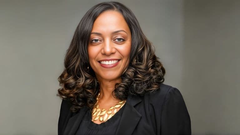 Delores Clemons, an African American woman with voluminous curls, wearing a black jacket and gold necklace, smiles confidently against a blurred gray background.