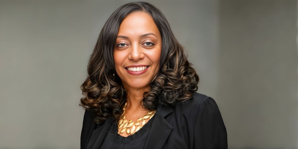 Delores Clemons, an African American woman with voluminous curls, wearing a black jacket and gold necklace, smiles confidently against a blurred gray background.
