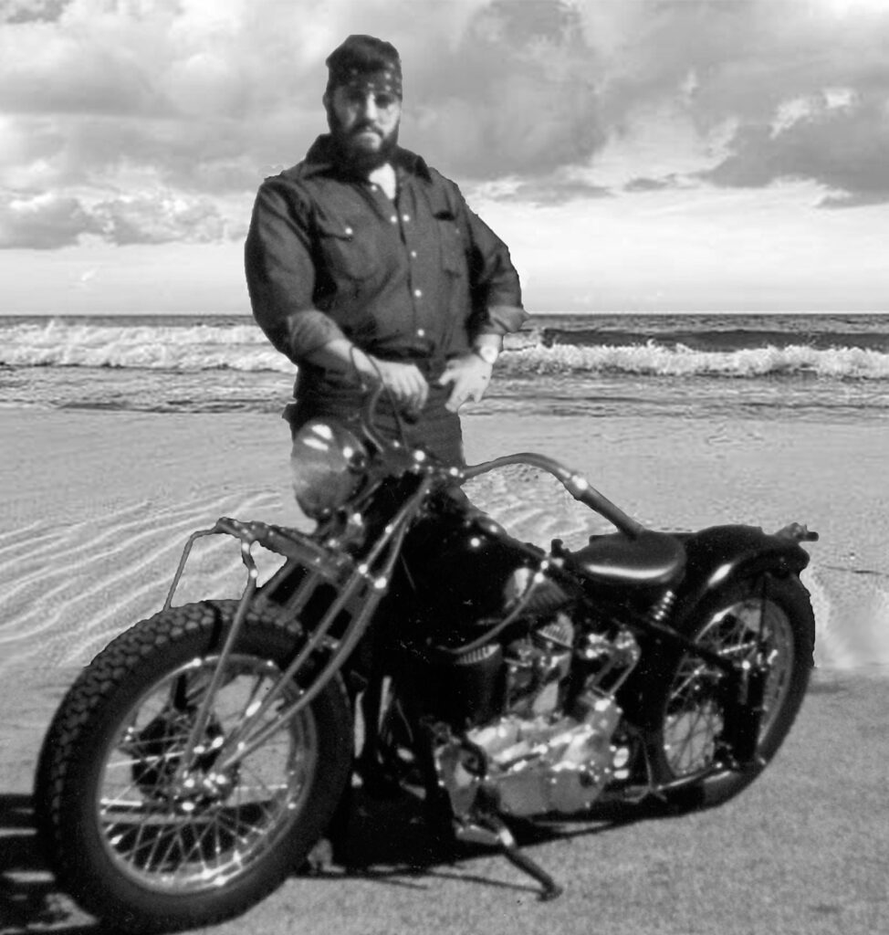 Vintage photo of Baer Charlton standing beside a custom motorcycle on a beach.